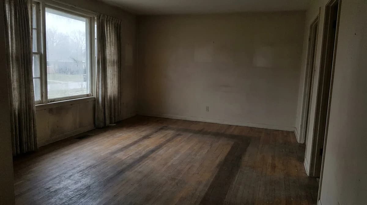 Foto inmobiliaria sin editar, apagada y subexpuesta, de un salón vacío con luz de ventana nublada
